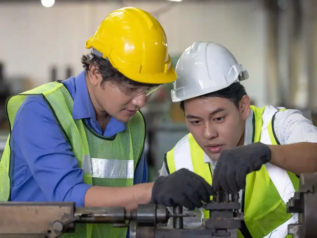 Two male industrial workers wearing safety vests, helmets, and gloves closely inspecting or assembling a machine part in a factory or workshop setting.