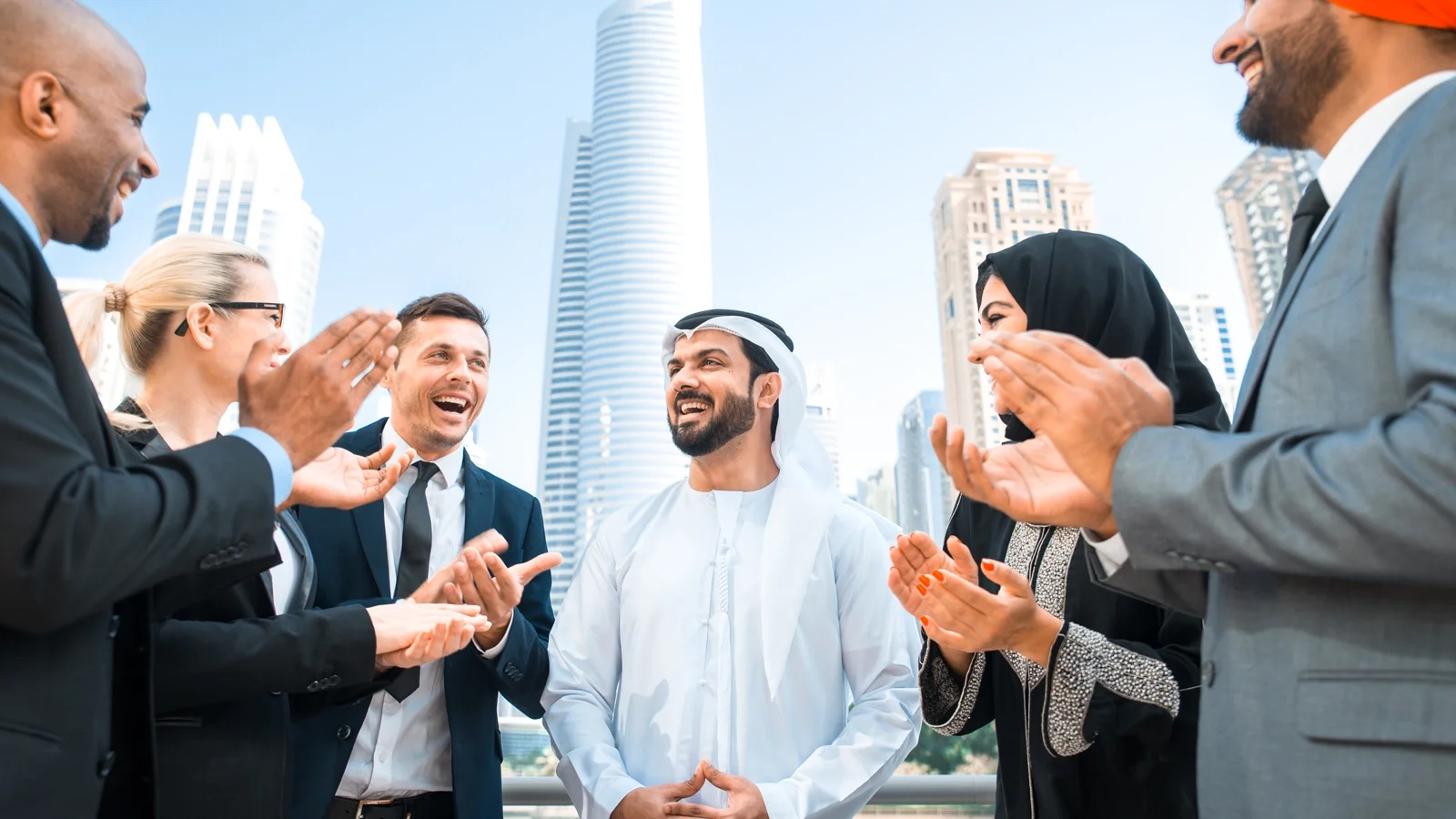 A group of business professionals stand outdoors in a modern cityscape, smiling and applauding. The man in the center wears traditional Middle Eastern attire, while the others wear business suits and formal clothing. Tall skyscrapers rise in the background under a bright, clear sky, creating a celebratory and collaborative atmosphere