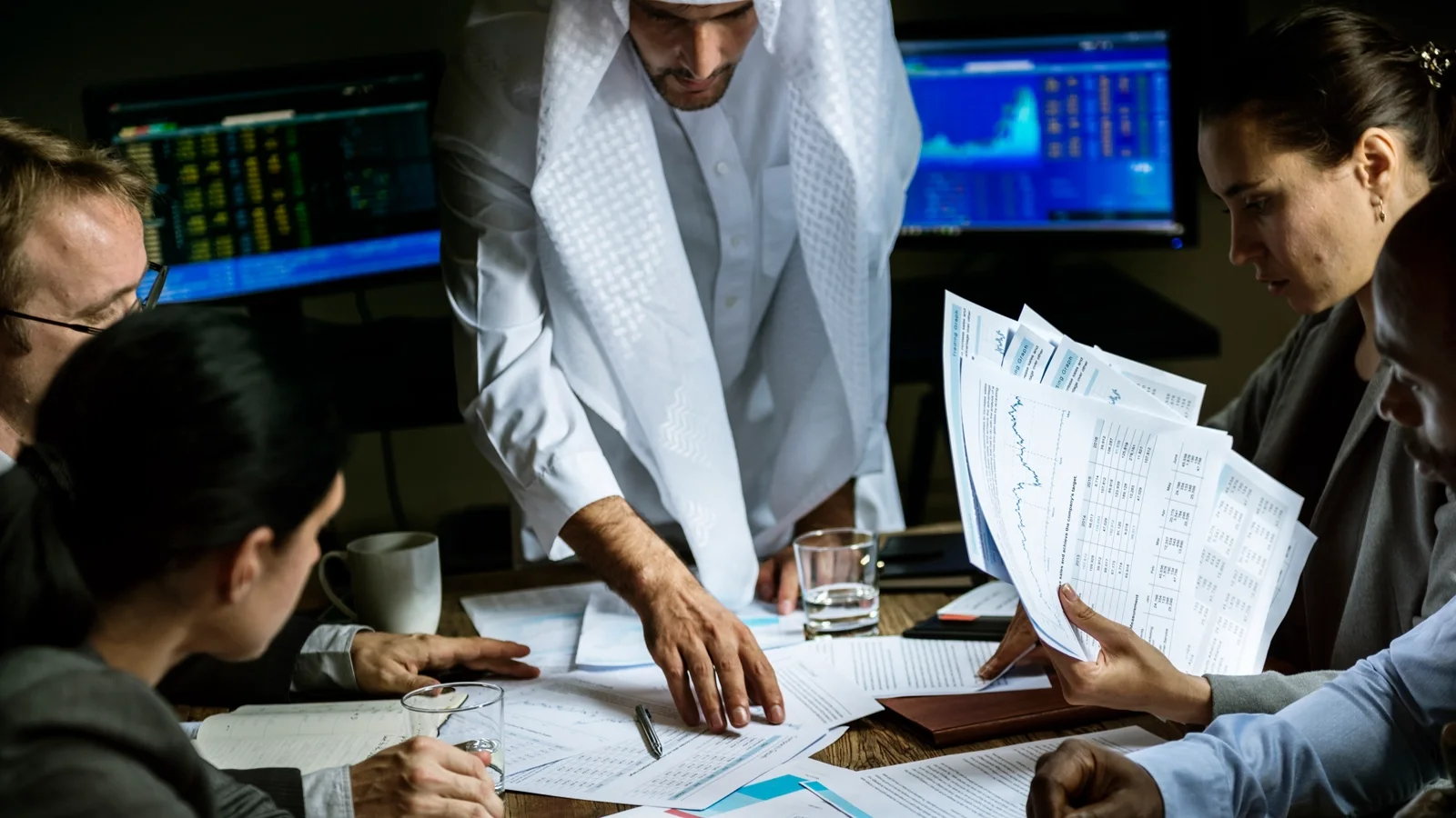 A group of business professionals sit around a table covered with documents, financial charts, and laptops. A man in traditional Middle Eastern attire leans over the table pointing at some papers, while others review printed financial reports. Computer monitors in the background display stock market data and graphs, suggesting an intense financial or strategic meeting