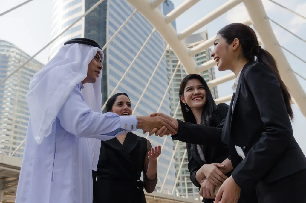 A group of business professionals standing outdoors in a modern city setting. A man wearing traditional Middle Eastern attire shakes hands with a woman in a business suit while two other women stand beside them smiling.