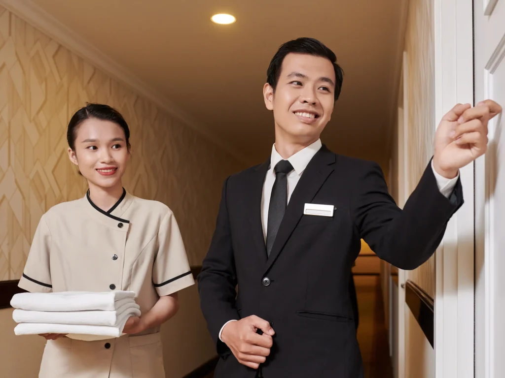 Dwóch pracowników hotelu na korytarzu, jeden w beżowym mundurku trzymający złożone białe ręczniki, drugi w czarnym garniturze z identyfikatorem wskazujący w stronę drzwi, wzorzysta tapeta i ciepłe oświetlenie.